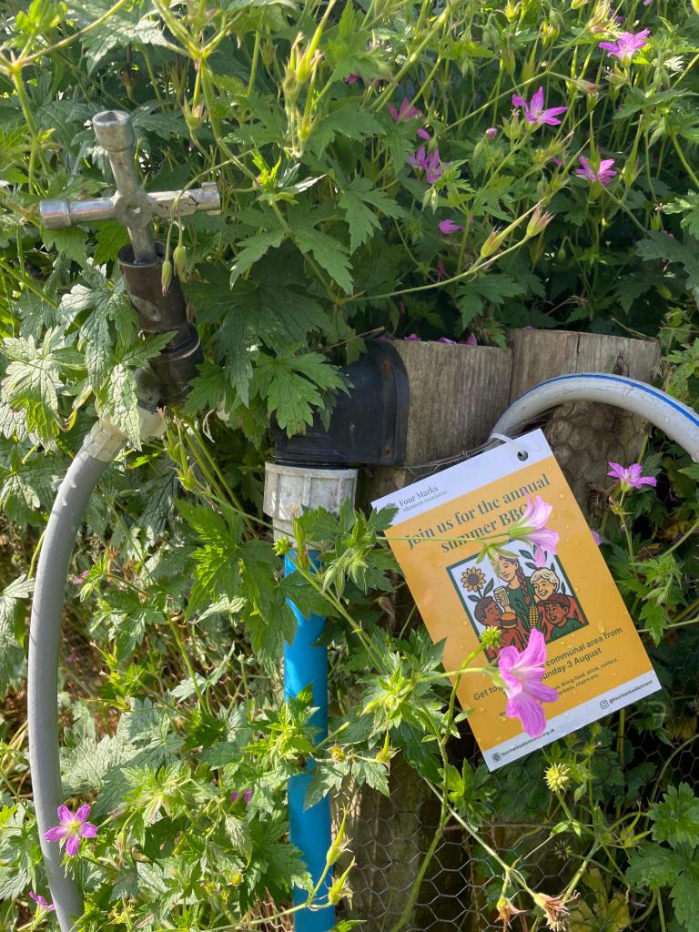 An FMAA summer BBQ flyer pinned to a communal tap, surrounded by pink geraniums and foliage.